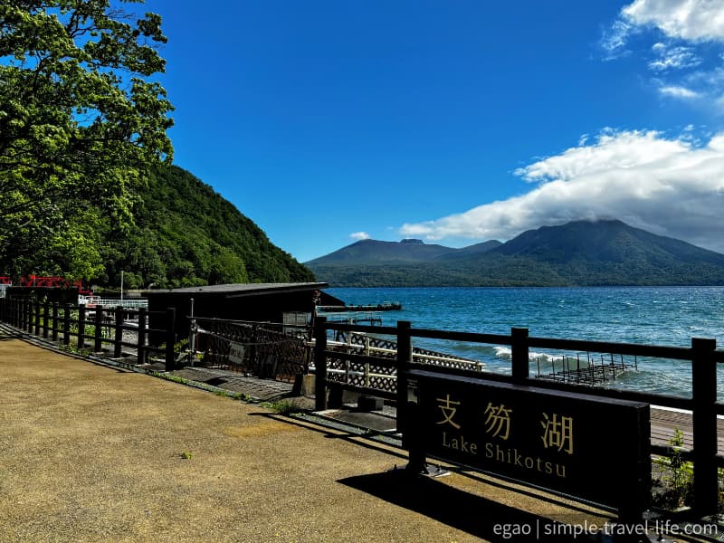 「支笏湖 Lake Shikotsu」の木の看板が立つ湖畔広場と、対岸に連なる山並み