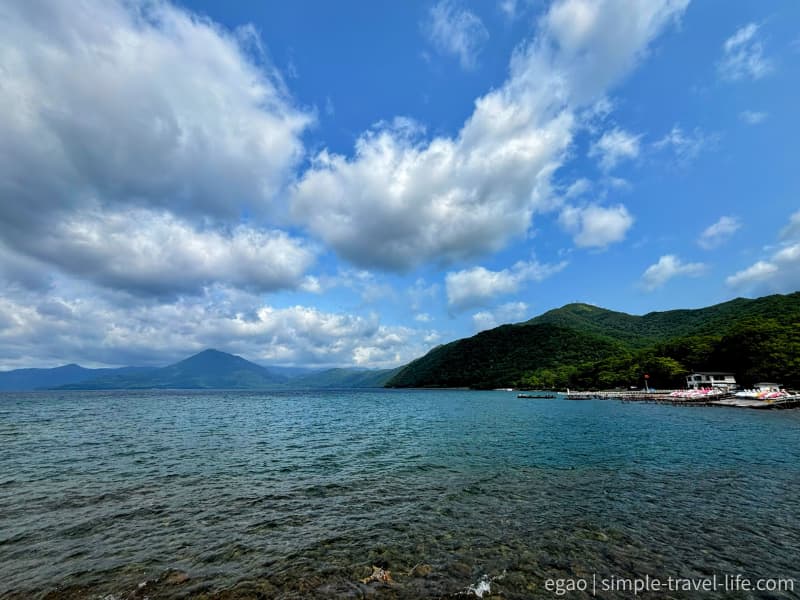 浅瀬まで透けて見える支笏湖の水面と、対岸の山並み、右手に桟橋とボート
