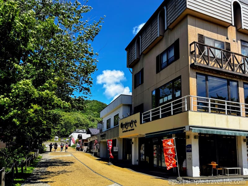 木立に囲まれた支笏湖温泉街の未舗装の通りと、通り沿いの店舗や赤いのぼり