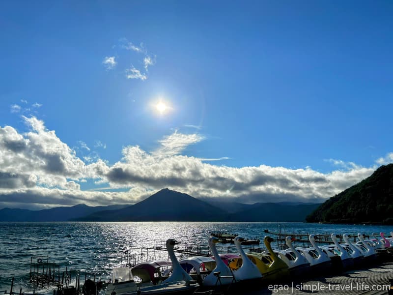 逆光に輝く支笏湖の湖面と、岸に並ぶ白鳥型のボート、対岸の山