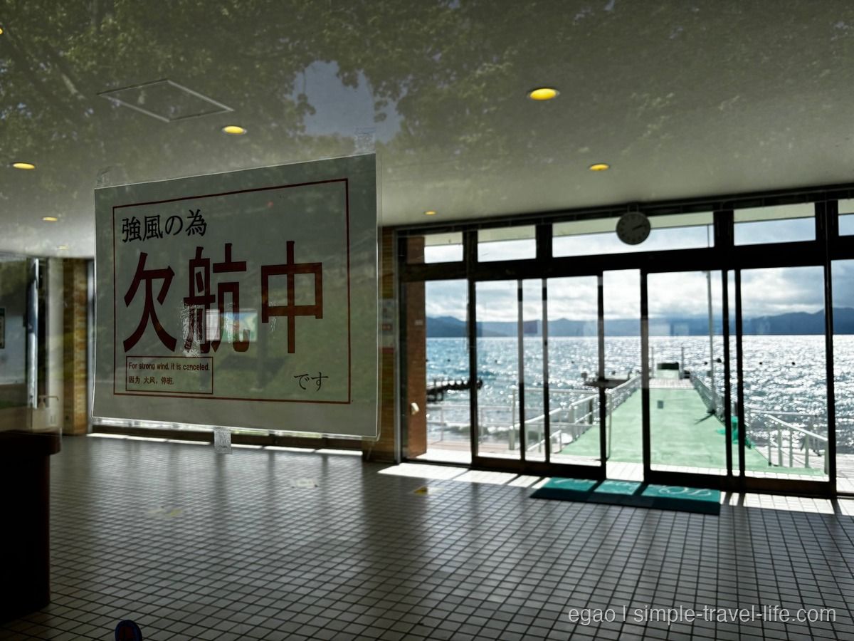 「強風の為 欠航中です」と書かれた掲示と、ガラス越しに見える桟橋と支笏湖