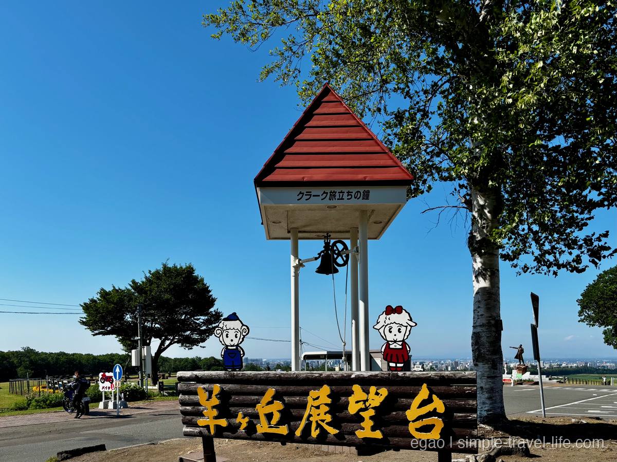「羊ヶ丘展望台」の丸太看板と、赤い屋根の「クラーク旅立ちの鐘」、羊のキャラクター2体