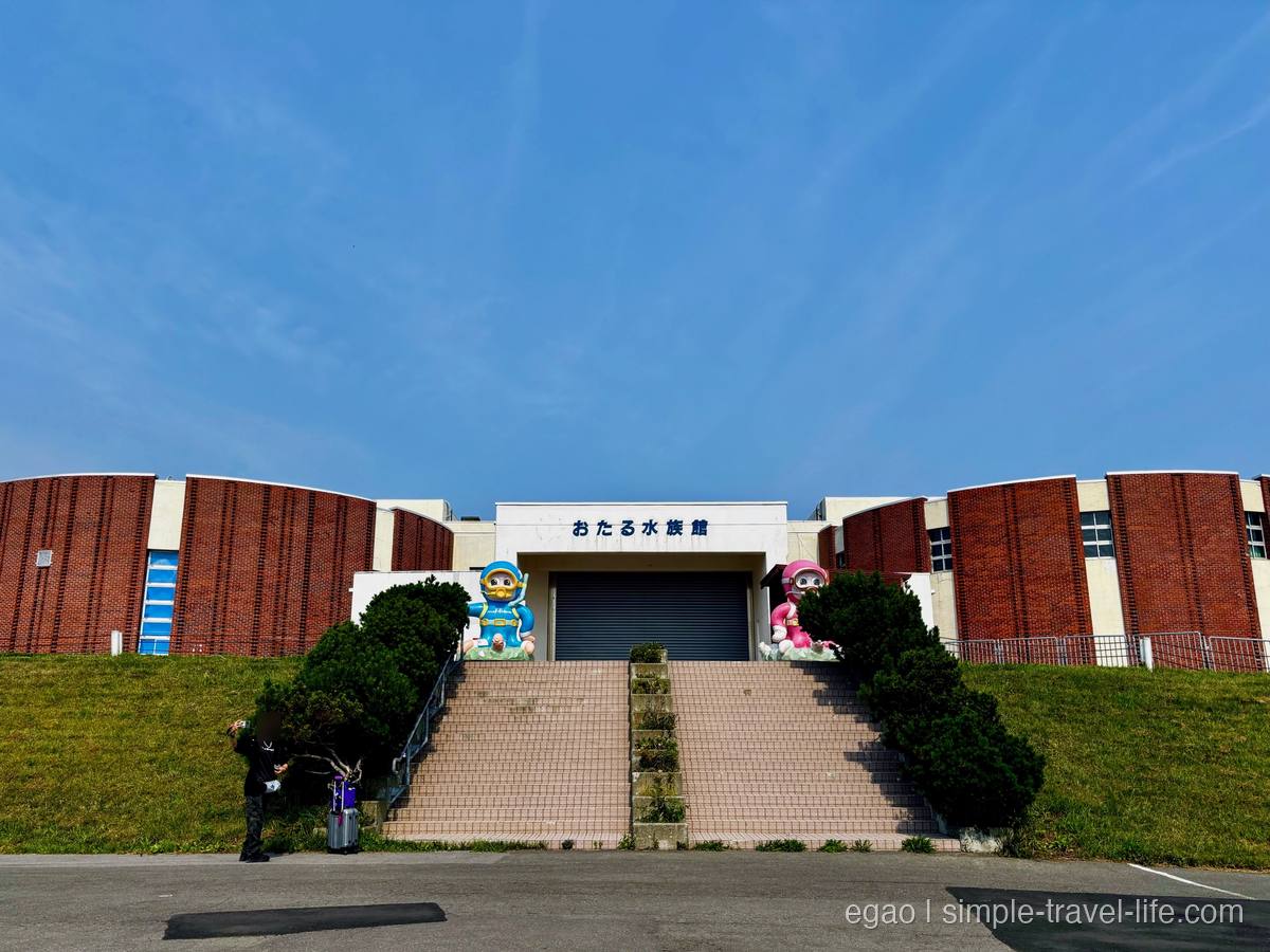 レンガ色の建物に「おたる水族館」の文字が掲げられた入口と、階段の両脇に立つダイバー姿のキャラクター像