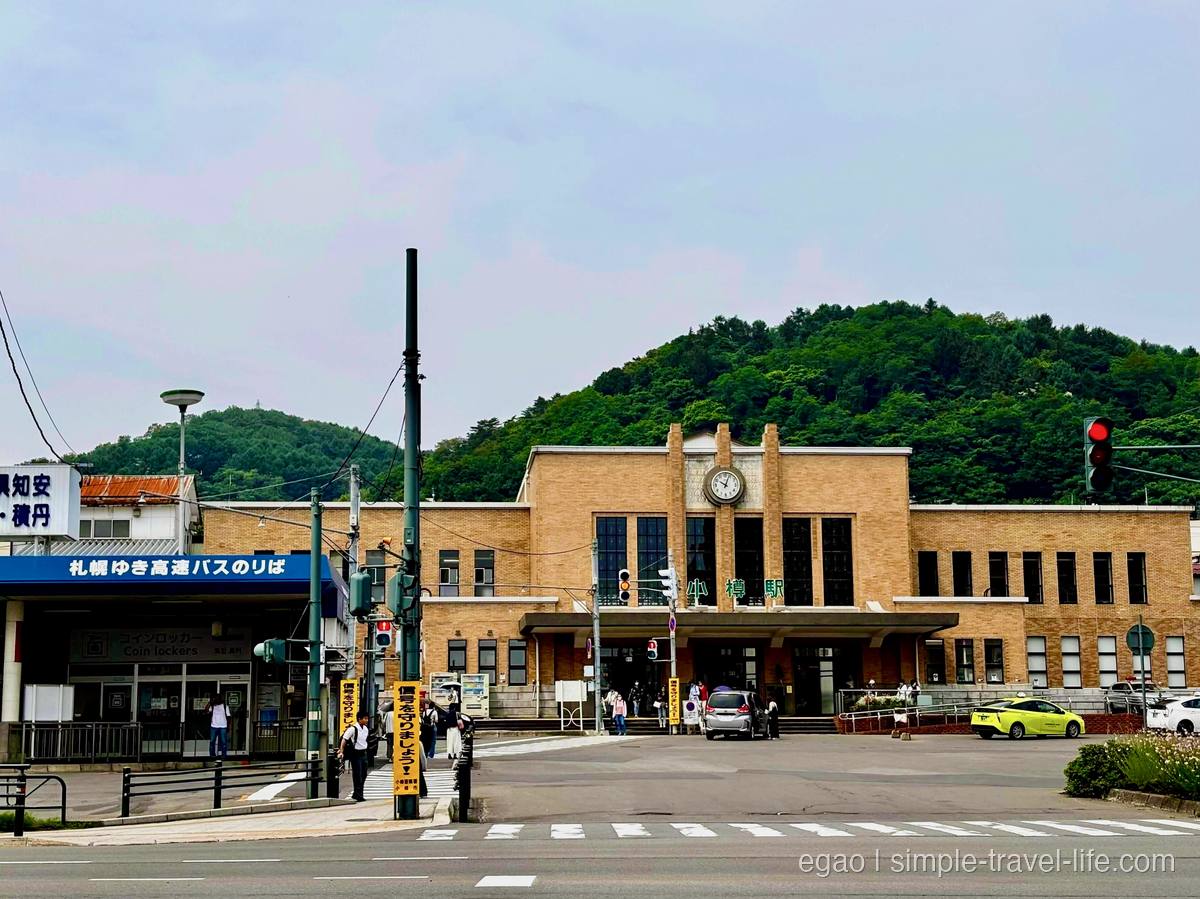 中央に時計を掲げた小樽駅の駅舎と、駅前に停まるタクシー、緑の山を背にした駅前広場