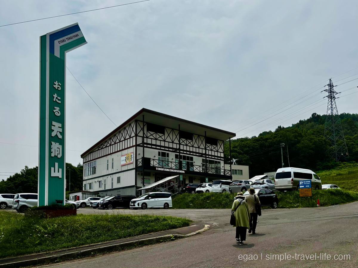 「おたる天狗山」と書かれた縦長の看板と、山麓の駐車場に立つ白と黒の三階建ての建物