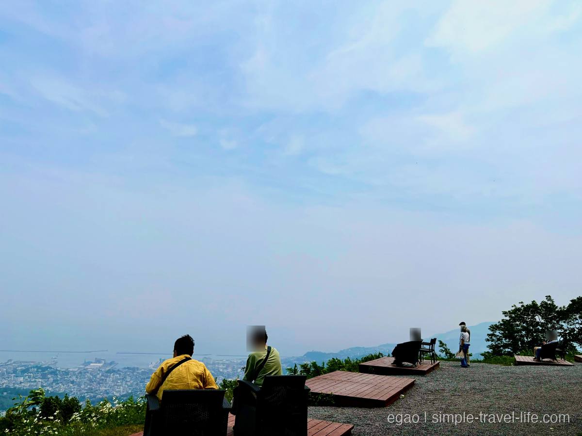 ウッドデッキの上に椅子が並ぶ天狗山山頂の展望スペースと、その先に広がる小樽の街と港