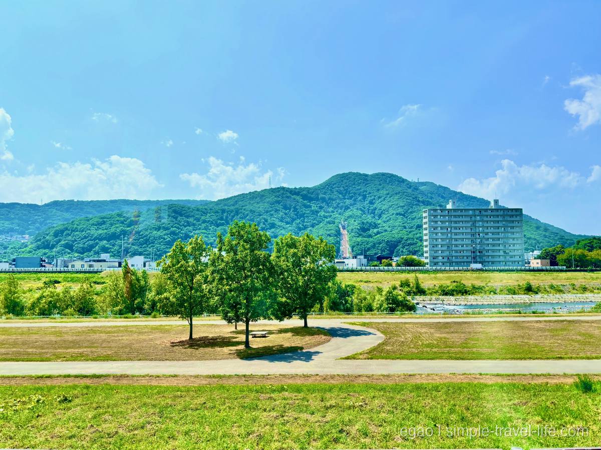 バスの車窓越しに見える、芝生の広い河川敷と遊歩道、その奥に建つ集合住宅と緑の山並み