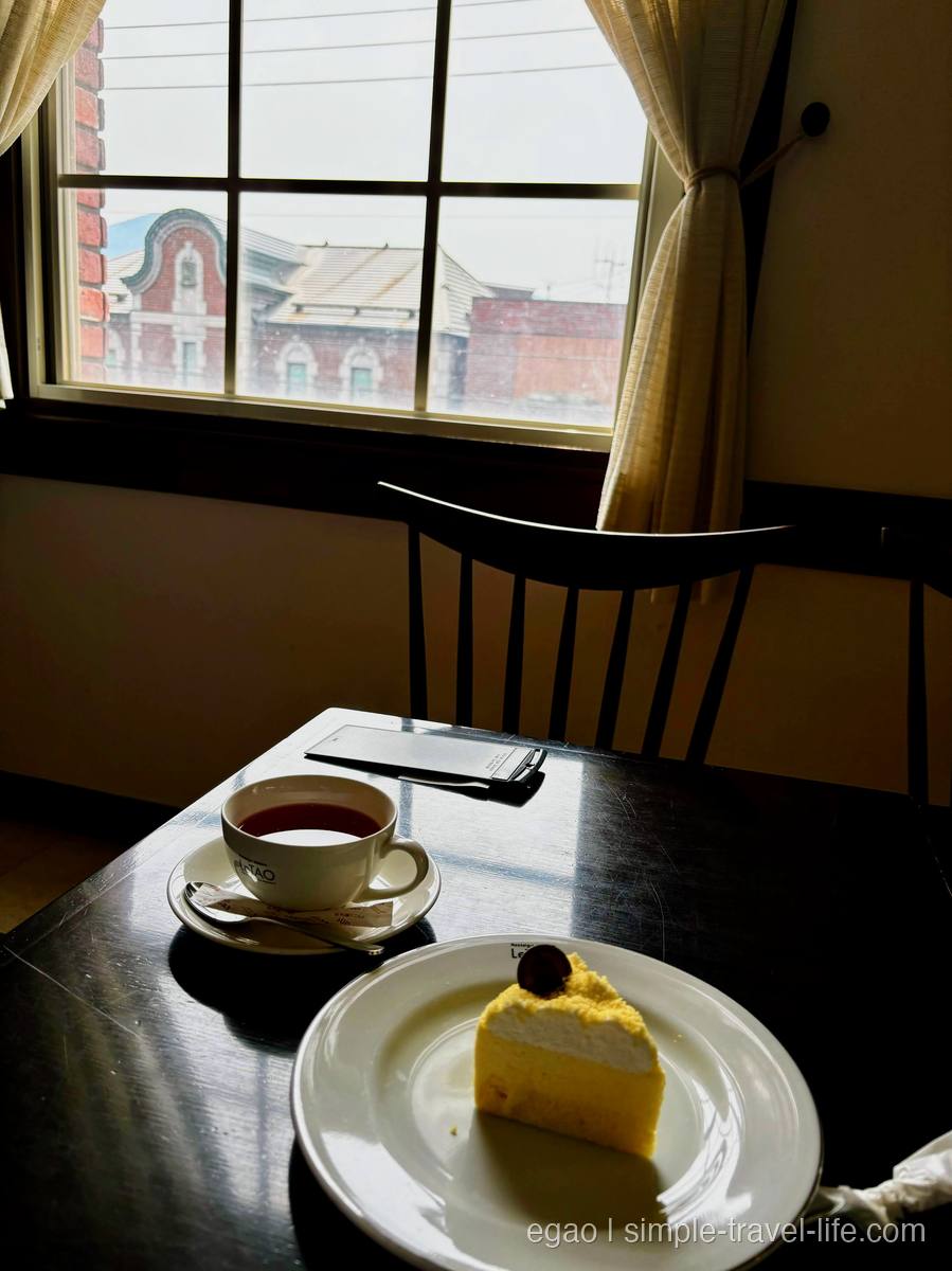 カフェの窓際のテーブルに置かれた紅茶のカップと、白と黄色の二層のケーキ。窓の外にレンガ造りの洋館