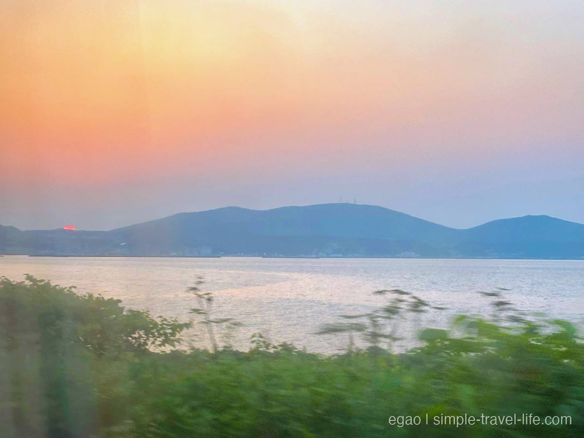 列車の窓越しに見える石狩湾の水面と対岸の山並み、山の端に沈みかけた夕日