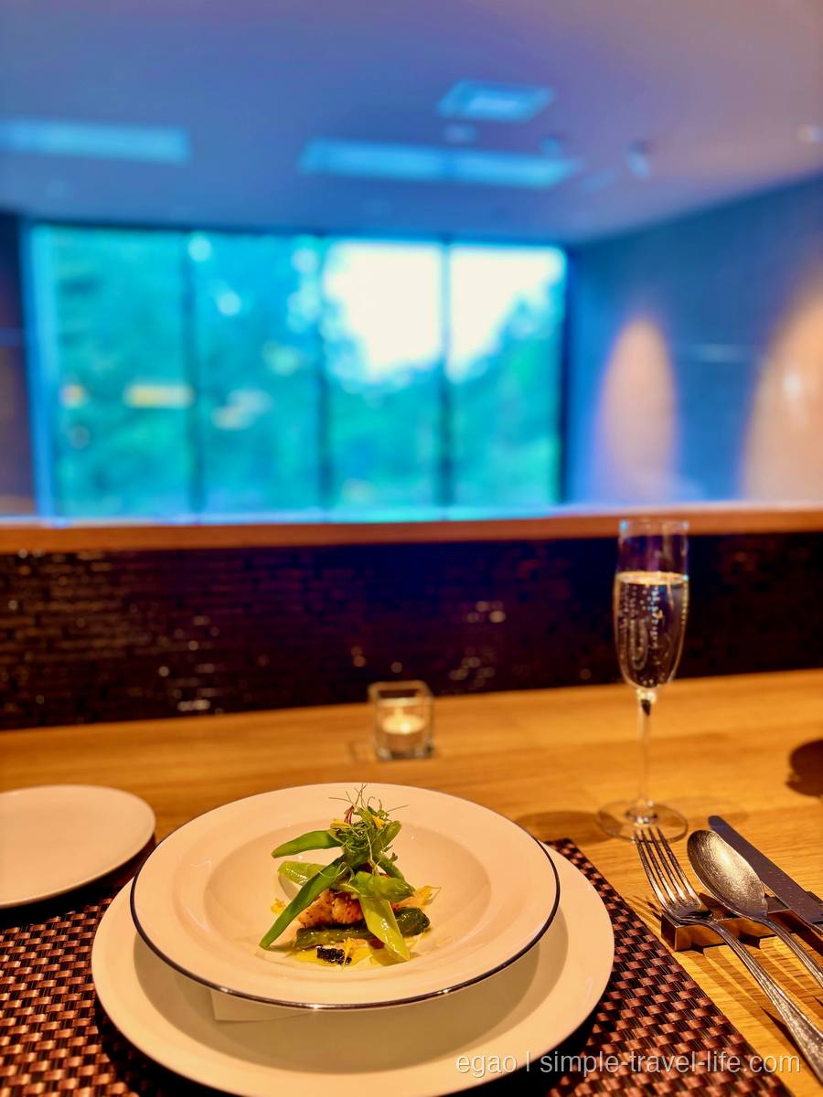 白い縁付きの皿に盛られたカニの身と絹さやなどの緑の野菜。奥にスパークリングワインのグラスとキャンドル