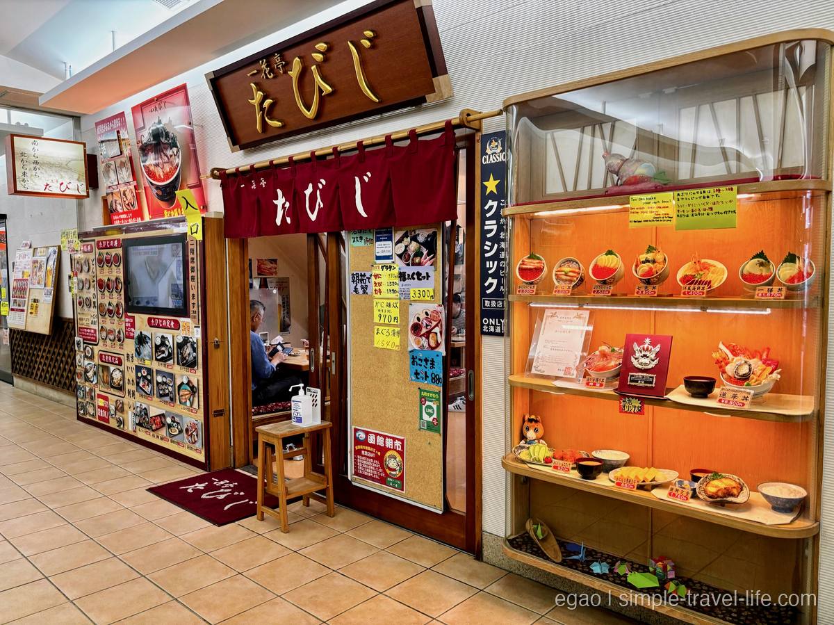 どんぶり横丁市場に並ぶ「一花亭たびじ」の赤いのれんと、丼の食品サンプルのショーケース