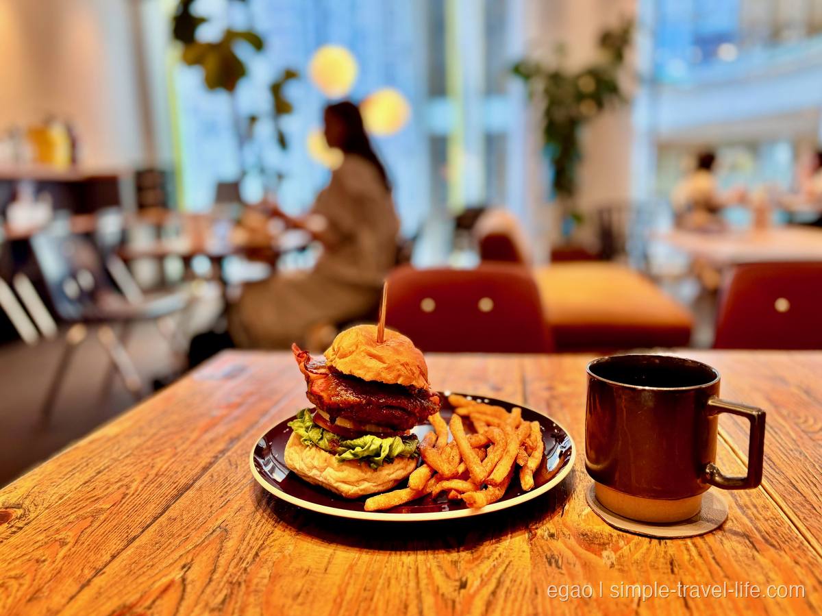 厚いパティとベーコンをはさんだハンバーガーとフライドポテトが載った黒い皿、隣にマグカップ