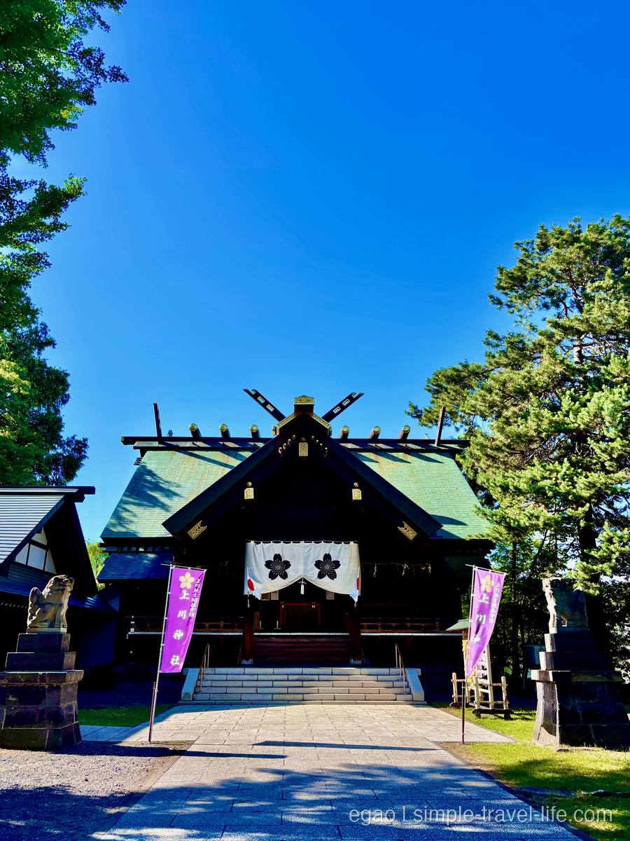 緑青色の屋根の木造の社殿と、「上川神社」と染め抜かれた紫ののぼり。石段の両脇に狛犬