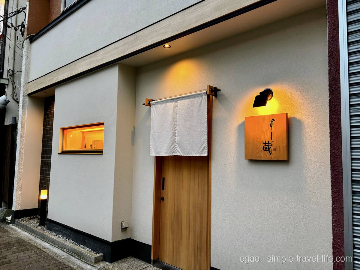 「すし蔵」の外観。白いのれんと木の引き戸、木の看板が照明に照らされている