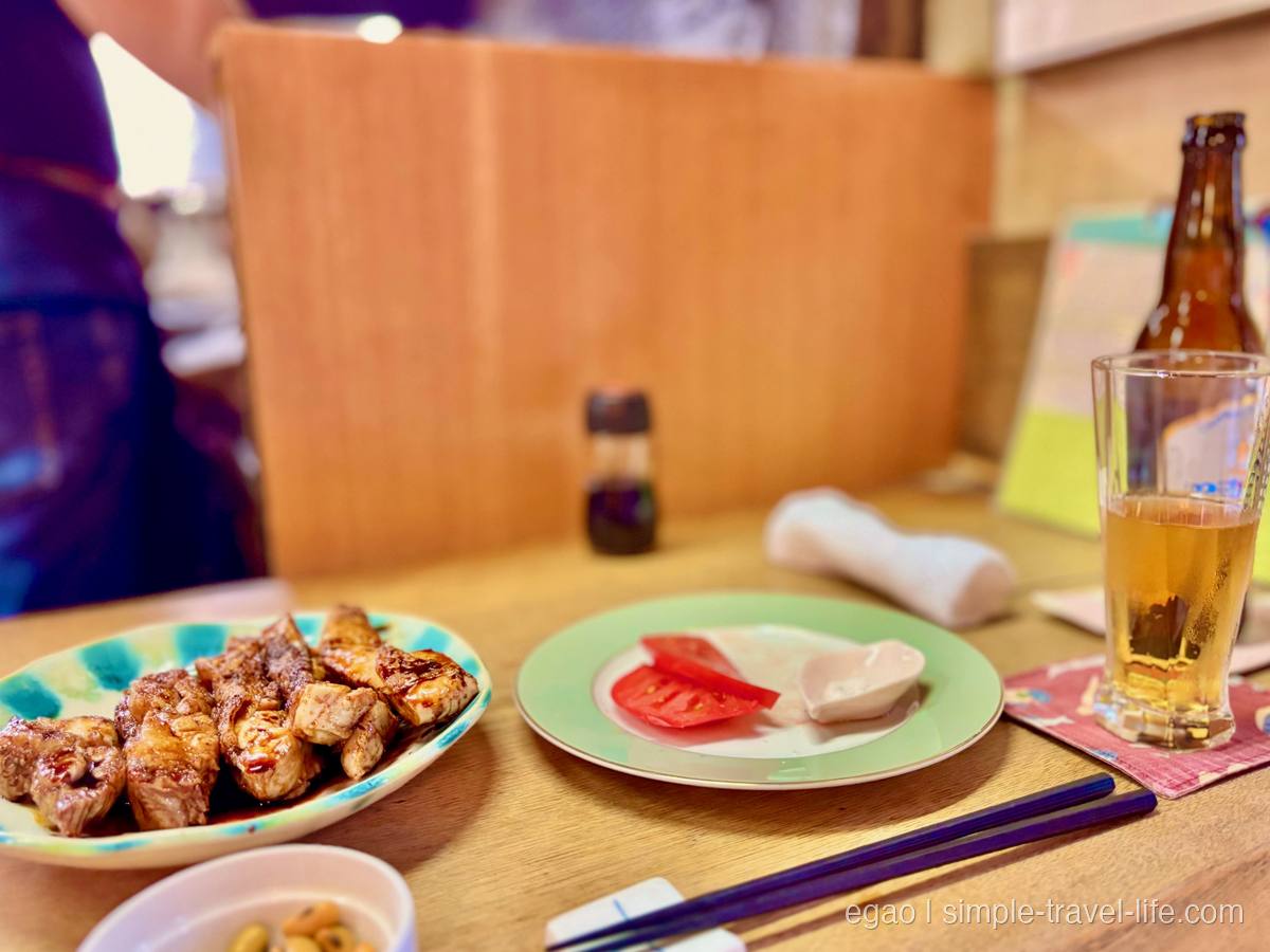 タレがかかった焼き鶏の切り身が並ぶ楕円の絵皿。奥にトマトを添えた皿と瓶ビール、グラス