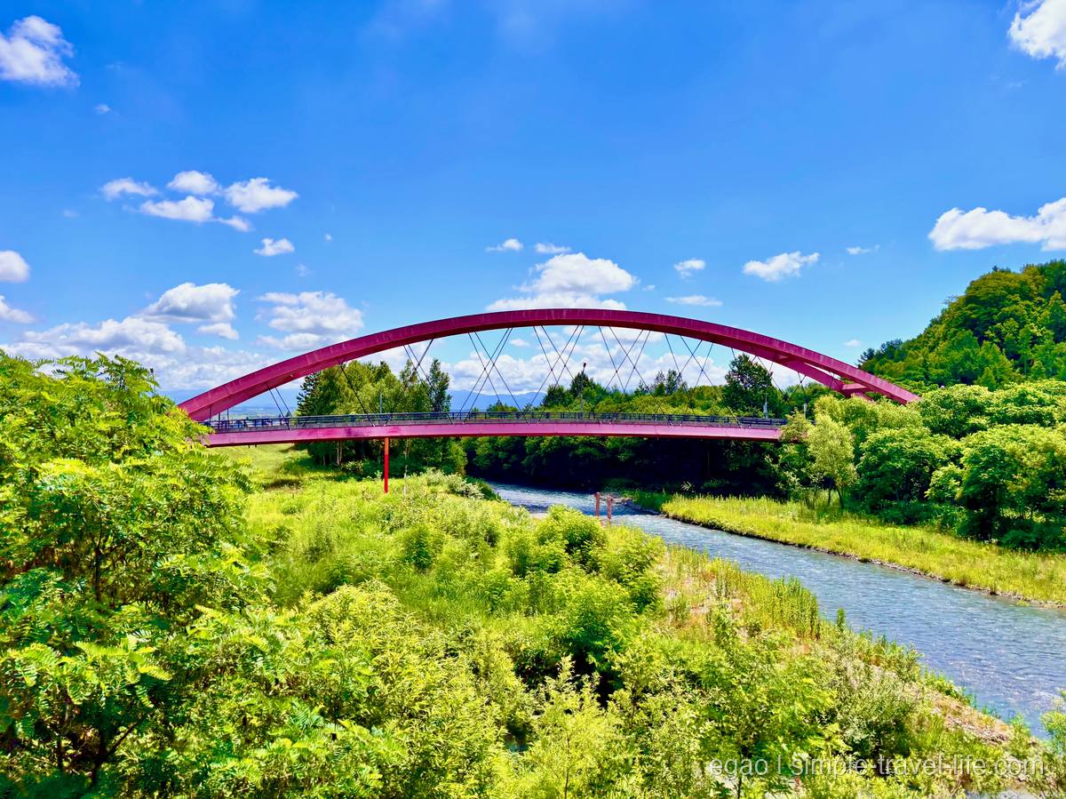 緑の木々にはさまれた川と、そこに架かる細い赤いアーチ橋。青空に白い雲が点々と浮かぶ