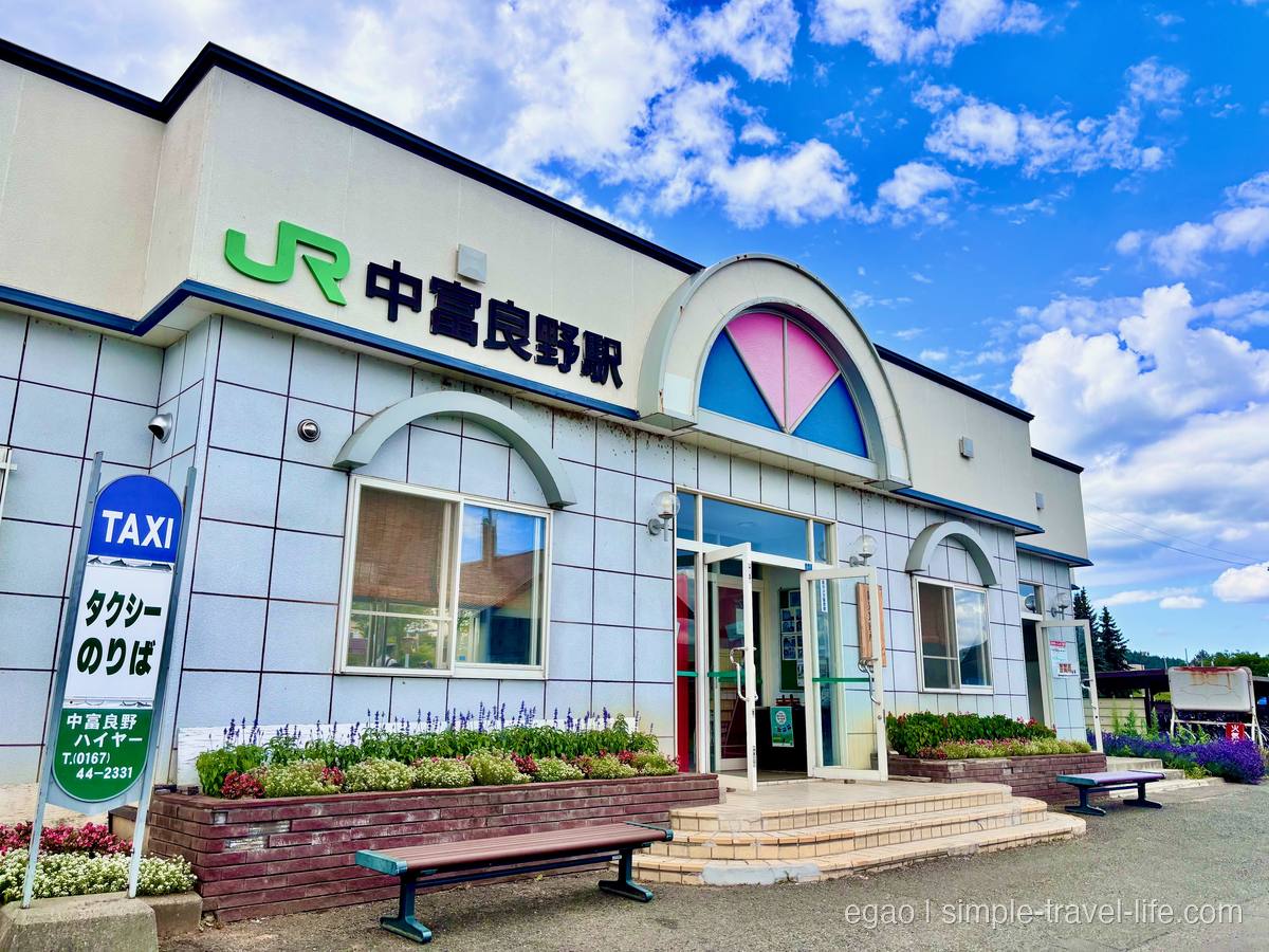 「JR 中富良野駅」と掲げられた白い駅舎。入口の上に半円の色ガラス、手前にタクシーのりばの看板とベンチ、青空