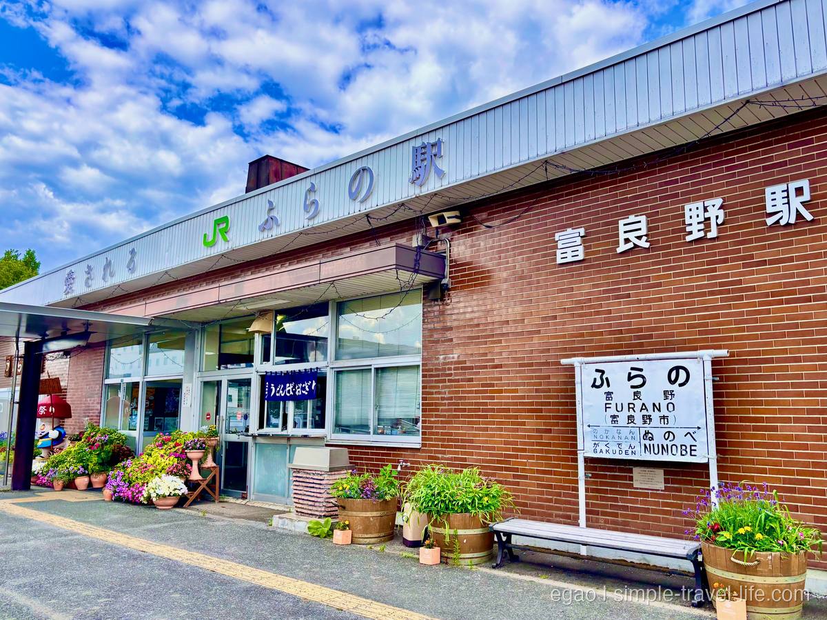 赤レンガと白い壁の富良野駅舎。「ふらの/FURANO」の駅名標と、花を植えた木の樽がいくつも並ぶ駅前