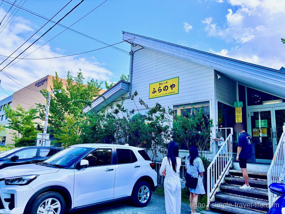 「カレーの ふらのや」の黄色い看板を掲げた白い建物。入口の階段に順番を待つ数人、手前の駐車場に車