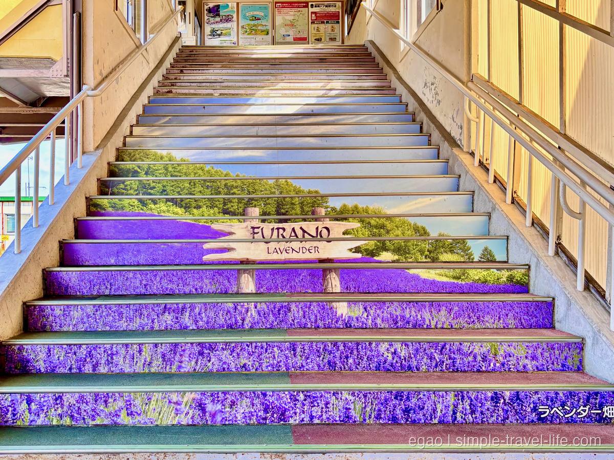 踏み面いっぱいにラベンダー畑の写真が貼られた駅の階段。中ほどに「FURANO LAVENDER」の木の看板、上の壁にポスター