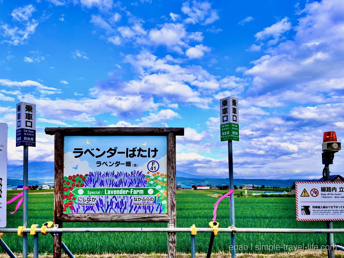 木枠の駅名標に「ラベンダーばたけ」の文字とラベンダーの絵。両脇に「乗車口」の標識、奥に緑の田んぼ