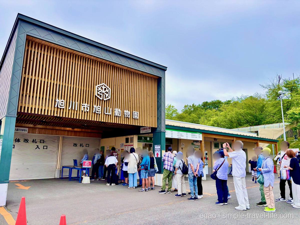 「旭川市旭山動物園」と掲げた木組みの入園ゲート。改札口の前に並ぶ大勢の人、うすい曇り空