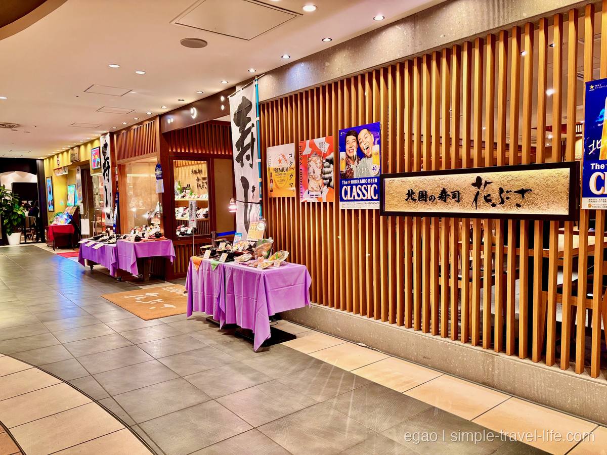 木の格子壁に「北国の寿司 花ぶさ」の木製看板。手前に紫の布をかけた台と料理見本、奥に「寿司」ののぼり