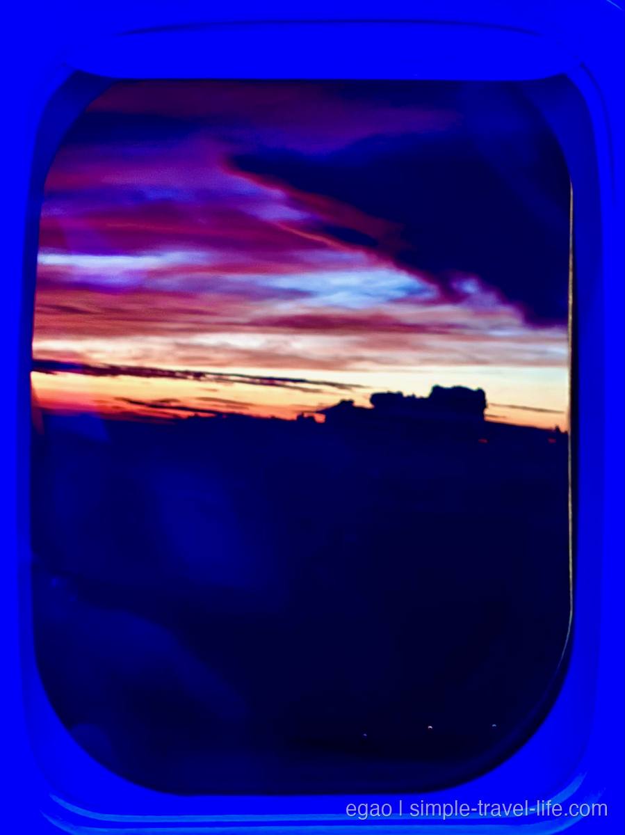 飛行機の窓の外に広がる夕焼け。紫とオレンジに染まった空と黒い雲、下側は暗い地上