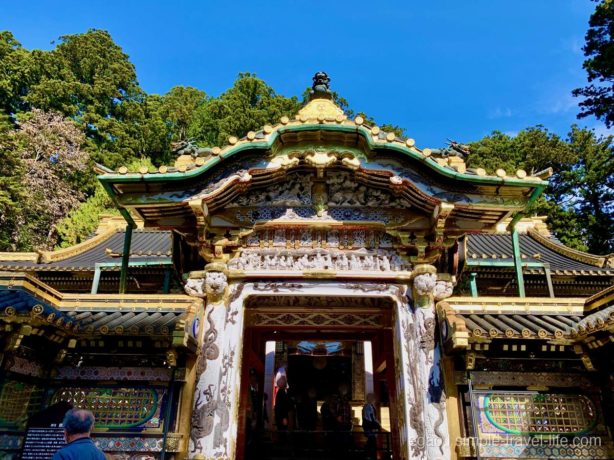 白い彫刻と金色の飾りで覆われた門を見上げた様子。屋根は緑と金、背後は杉の木と青空
