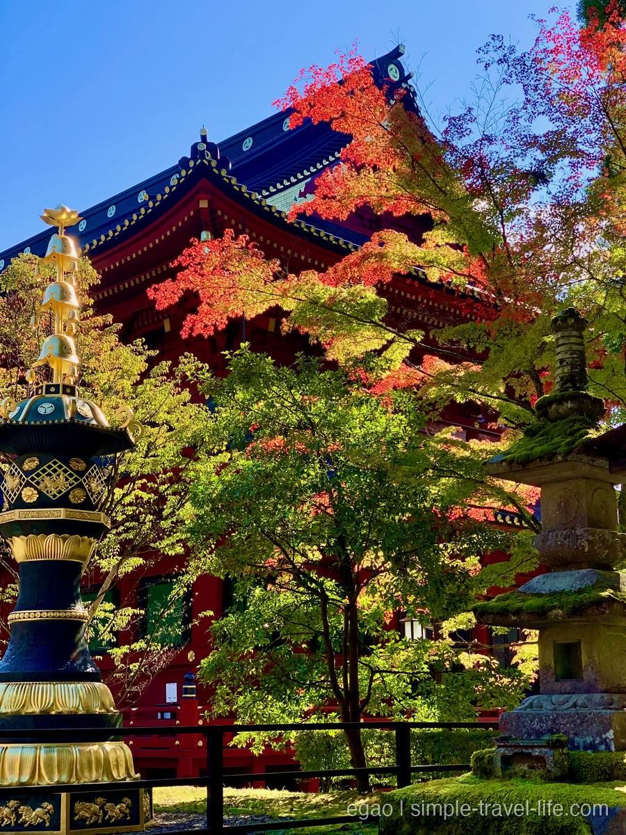 輪王寺の境内に立つ大きな青銅の灯籠。金色の宝珠と鈴が段に重なり、笠に三つ巴の紋。右手前に苔むした石灯籠、奥に朱塗りのお堂の屋根と、赤や黄の紅葉